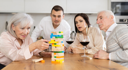 Cheerful senior woman spending free time with husband and adult children at laid-back family home get-together with drinks, concentrating on removing block from tower while playing jenga