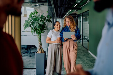 Two business women standing in an office corridor collaborating and discussing work using a digital...