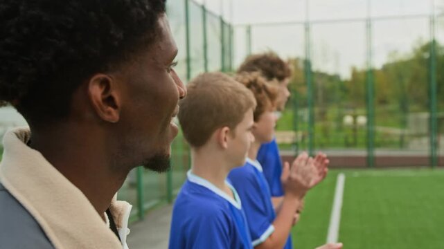 Close up of young African American coach and three bench players smiling, clapping and screaming to cheer up their team during football match