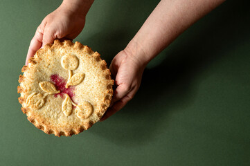 Hands holding a round baked pie with a red filling detail in the center on a green surface.