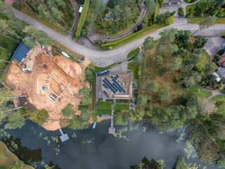 Aerial view of a modern house with solar panels near a water body with docks, adjacent to a construction site, surrounded by trees and gardens.