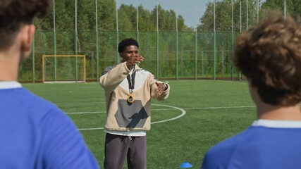 Back view shot of three football players standing on pitch, looking at African American coach explaining game strategy