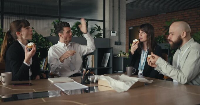 Four colleagues eating pastries and chatting together during a casual office break in a modern workspace with a relaxed atmosphere and friendly vibes