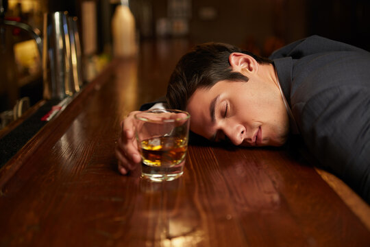 Man in suit lies forehead on bar beside whiskey glass, capturing inebriation and exhaustion in dimly lit pub scene.