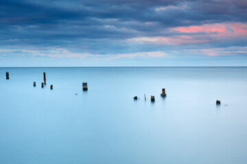 A calm blue ocean with a few wooden posts floating in it. The water is still and the sky is cloudy