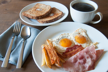 Fried eggs with ham and French fries on a white plate served as breakfast with black coffee and toast with butter on a wooden table with a 45-degree viewing angle