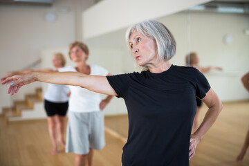 Active mature woman visiting choreography class with group of aged females, learning classical ballet technique .