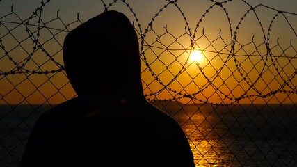 Refugee at sunset near the border. Hooded silhouette standing near barbed wire fence, gazing at golden sunset horizon, symbolizing hope, freedom, and inner struggle amid confinement and isolation - Powered by Adobe