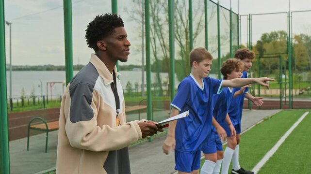 Serious African American coach and three bench players watching football match on open air pitch