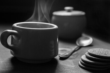 Steaming cup of coffee, with cookies on one side and a sugar bowl with a spoon on the bottom, on a wooden table against a black background. Still life black and white photography.