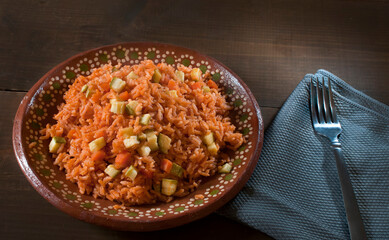 Mexican rice on a clay plate with a fork next to it on a rustic wooden table, with a blue cloth napkin, top view lay flat
