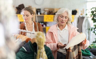 Mature woman customer examine and choose plaid jacket in store, and buyer consider some items. Concept of seasonal wardrobe shopping. Client enjoys shopping for stylish clothes