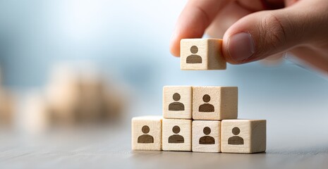 A hand carefully places a small wooden block, featuring a person icon, atop a small stack of similar blocks.  The blocks form a rudimentary hierarchy