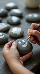 A close-up view of a child's hands carefully painting a white smiley face onto a smooth grey stone with a small brush during a creative craft activity.
