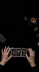 A person's hands are carefully arranging dark, mystical tarot cards on a black surface, surrounded by glowing crystals and a flickering candle, viewed from directly above.
