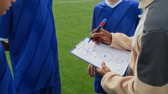 Over shoulder view of unrecognizable coach drawing game tactic on paper while football players standing near