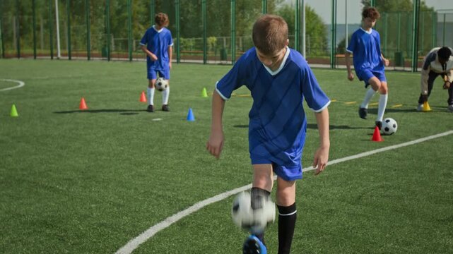 Preteen boy in blue kit juggling football and catching it, his teammates kicking football in background