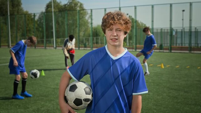 Slow motion of curly haired preteen Caucasian boy holding football and squinting from sunrays while standing on pitch with his coach and teammates