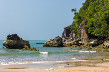Gentle waves washing ashore on a sandy beach beside jungle-covered rocks in Malaysia.