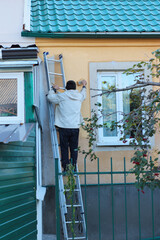 Man standing on a ladder painting the exterior wall of a house near the window. Outdoor renovation...
