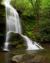 A stunning waterfall cascades down rocky cliffs, surrounded by vibrant green trees and plants. The water flows into a calm pool below, creating a peaceful atmosphere. Catawba Falls, North Carolina.