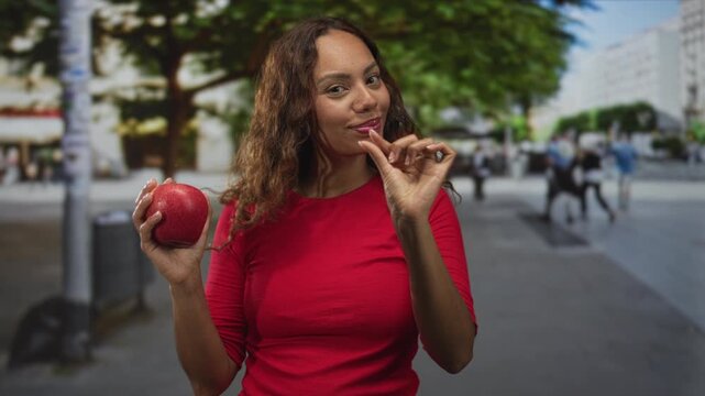 Woman holding a red apple and pinches her fingers to show a tiny bite or size on a city street plaza; healthy choice playful.
