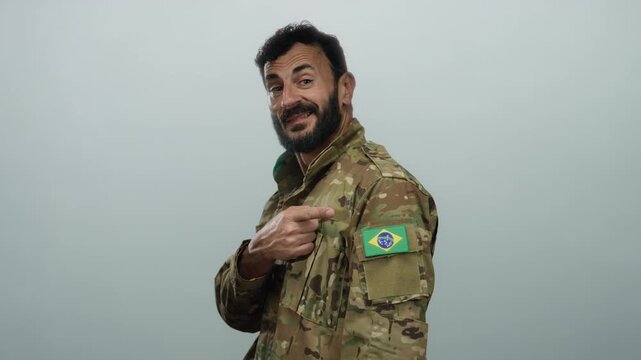 Hispanic man in military uniform points to brazilian flag patch on his sleeve, standing against a grey background with a proud expression and a beard.