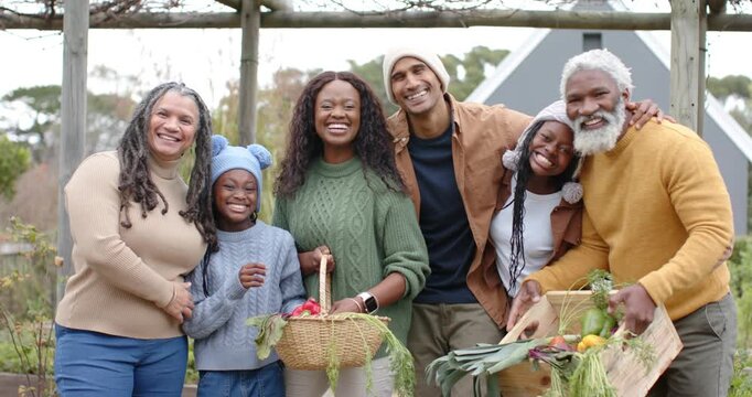 Posing under arbor, diverse family holding baskets, crate, girl lifting red pepper sharing harvest