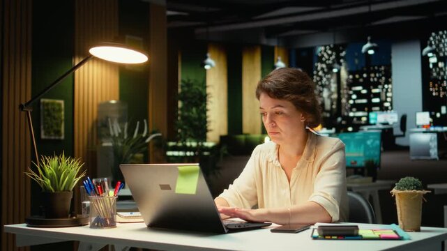 Weary businesswoman stays connected on social media while she does overtime at night, checking her mobile phone after receiving a notification. Female worker checks text messages. Camera A.