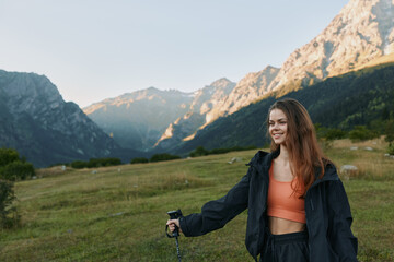 Fototapeta premium A cheerful woman hiker enjoys an outdoor moment in a mountain meadow. She smiles warmly, wearing a jacket and orange top, holding a trekking pole as sunlight highlights the alpine scenery.