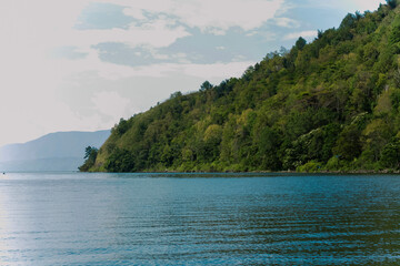 Naklejka premium Foto lanskap alam yang menampilkan keindahan danau dengan air biru jernih, dikelilingi perbukitan hijau yang rimbun. Di tepi danau terlihat rumah-rumah sederhana serta pepohonan tropis yang asri, menc
