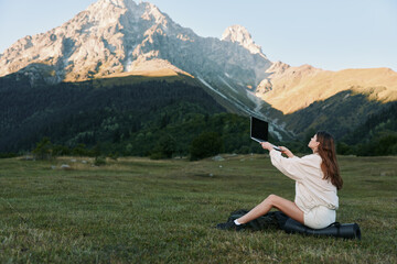 A woman sits on a small bag in a grassy field, using a laptop with a mountain backdrop. She appears...