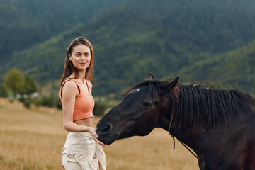 A confident woman stands beside a dark horse in an open field beneath distant mountains. She smiles...