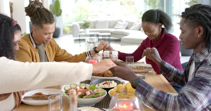 Diverse friends reaching across table at home, holding hands, bowing heads in prayer before meal