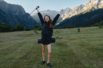 Fototapeta premium A cheerful hiker stands in a grassy valley with towering mountains in the background, raising a trekking pole and smiling, enjoying an adventurous outdoor moment.