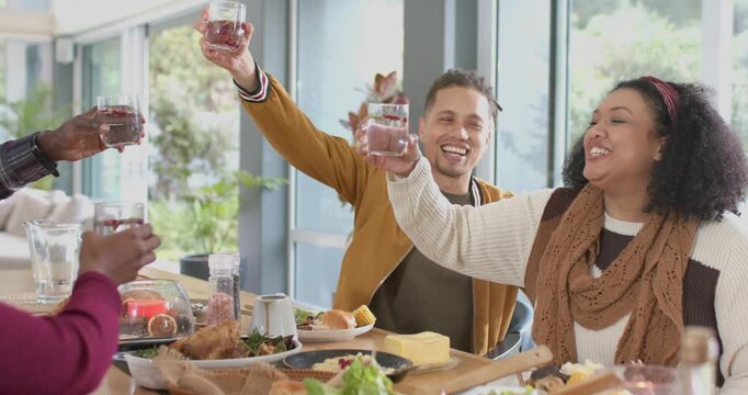 Diverse friends clinking glass tumblers, man raising drink at home dining table for brunch toast