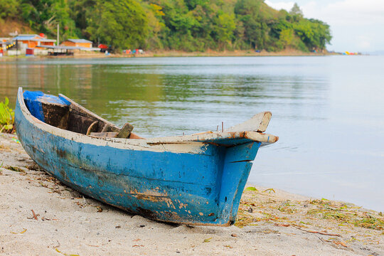 Foto perahu kayu biru yang terdampar di tepi danau dengan suasana perkemahan di latar belakang. Terlihat tenda berwarna-warni, beberapa sepeda motor, serta pepohonan hijau yang menambah nuansa alami d