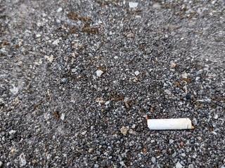 A close-up of a coarse, gray gravel surface scattered with small stones and dirt. A discarded white cigarette butt is visible in the foreground, highlighting themes of urban pollution and litter.