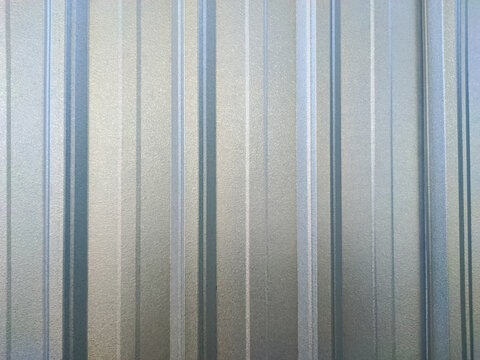 A corrugated galvanized steel surface, with a reflective, matte gray and silver finish. The texture features vertical ridges and subtle imperfections, creating an industrial, abstract background.