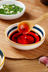 Red chili oil in wooden spoon dripping into bowl with fresh herbs and garlic on kitchen table