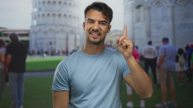 Young man smiling and pointing up at the iconic leaning tower of pisa, surrounded by people on a sunny day, capturing a moment of joy and wonder in italy's historic cityscape.