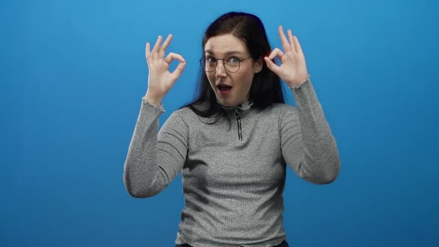 Woman with brunette hair makes an okay gesture smiling confidently against a vibrant blue background in a casual gray sweater and glasses.