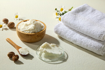 Wooden bowl of creamy shea butter with nuts, chamomile, glass dish and soft folded towel, shot on white surface for natural beauty visuals.