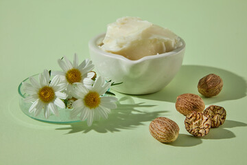 Small bowl of shea butter placed with chamomile flowers and nuts on green background. Clean minimalist setup for skincare product visuals.
