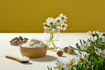 Shea butter in jar with chamomile bouquet, nuts and wooden spoon arranged against yellow backdrop, styled for cosmetic and spa advertising.