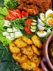 Close-up of Indonesian platter featuring corn fritters (perkedel jagung), fresh cucumber and lettuce, spicy red sambal served in banana-leaf bowl, boiled eggs and shredded fried chicken