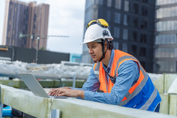 Asian construction engineer wearing safety vest and helmet working on laptop at construction site,...