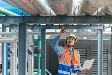Asian industrial engineer wearing safety gear inspects HVAC system with walkie-talkie and laptop, monitoring pipeline and equipment operation at rooftop facility for maintenance and safety.