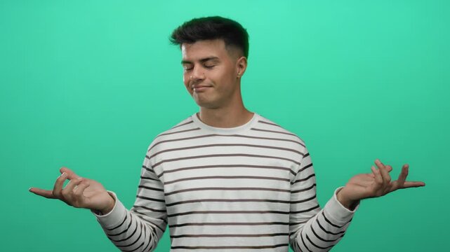 Young man in striped shirt gestures over isolated green background, looking puzzled and confident, expressing indifference and calm, conveying a relaxed mood.