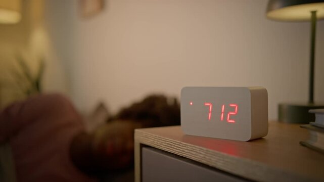 Medium selective focus shot of sleepy unrecognizable African American woman waking up in morning at home, stumbling out of bed and struggling to switch off beeping alarm clock